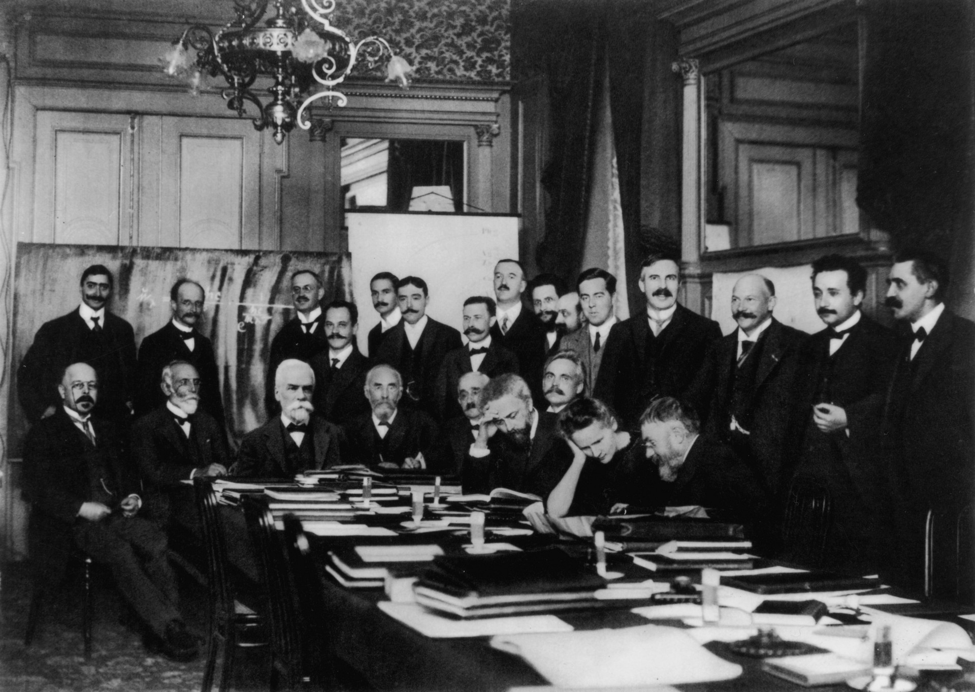 At First Solvay Conference (1911), Marie Curie (seated, second from right) confers with Henri Poincaré; standing, fourth from right, is Rutherford; second from right, Einstein; far right, Paul Langevin